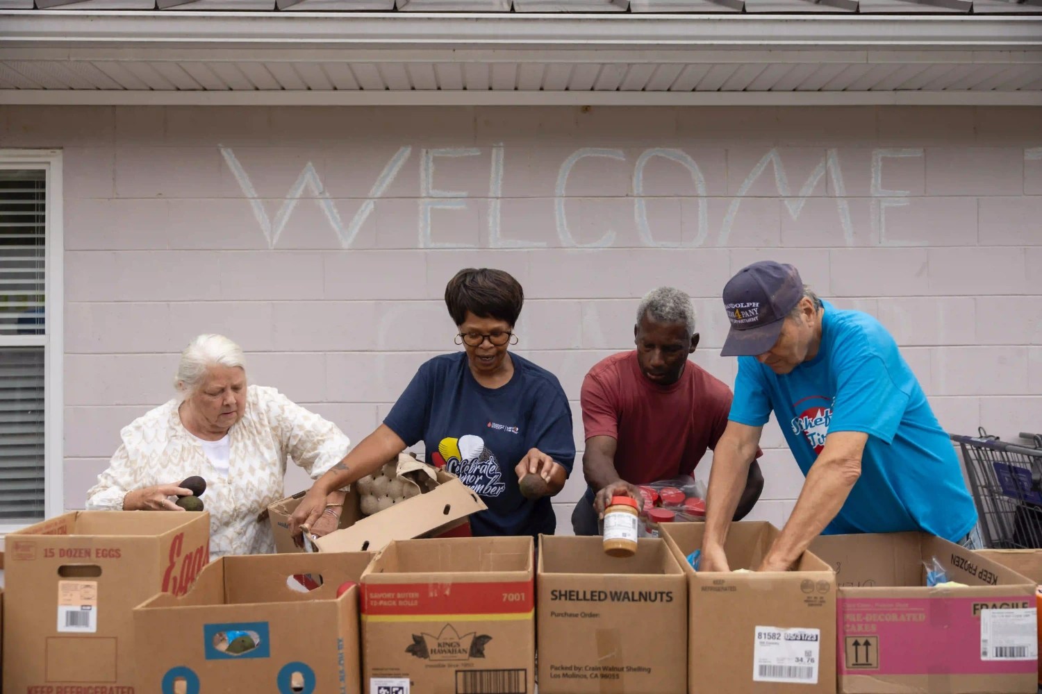 Four volunteers at Feed More sort food into different boxes, with a "Welcome" sign behind them.